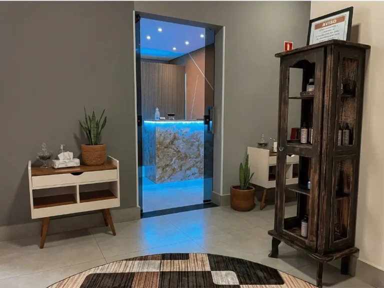 Visão interna da clínica da Dra. Lais Vieira, mostrando os detalhes da decoração premium. O ambiente apresenta um armário rústico em madeira escura exibindo produtos de saúde, um aparador em estilo vintage com plantas e um tapete geométrico, com vista através da porta de vidro para a recepção iluminada ao fundo.