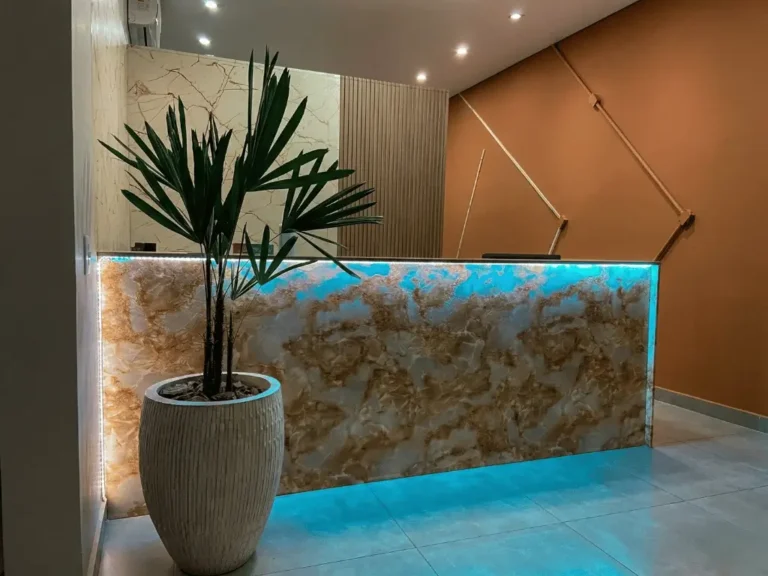 Balcão de recepção da clínica da Dra. Lais Vieira em Santa Bárbara d'Oeste. O design de interiores destaca um imponente balcão em pedra translúcida com iluminação embutida azul, contrastando com painéis ripados e uma parede terracota geométrica, transmitindo sofisticação e cuidado desde a entrada.