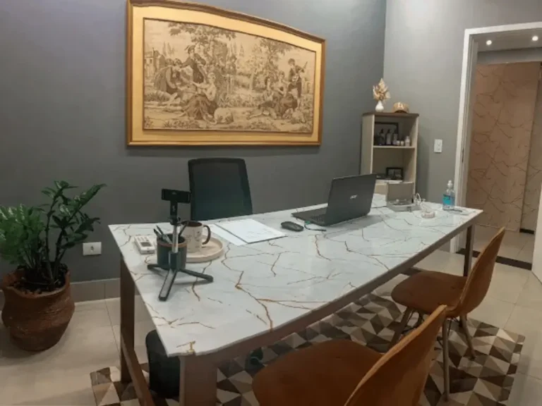 Visão da sala de atendimento da clínica de medicina integrativa da Dra. Lais Vieira. O ambiente possui uma ampla mesa com textura de mármore claro, cadeiras estofadas em tom caramelo, paredes em cinza sofisticado com um quadro clássico e decoração acolhedora, projetado para consultas exclusivas e sem pressa em Santa Bárbara d'Oeste.
